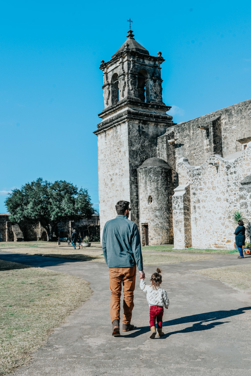 Top 10 Kid Friendly Activities in San Antonio by popular Texas travel blog, Lone Star Looking Glass: image of a a dad holding his 2 year old daughter's hand as they walk through Mission National Park.