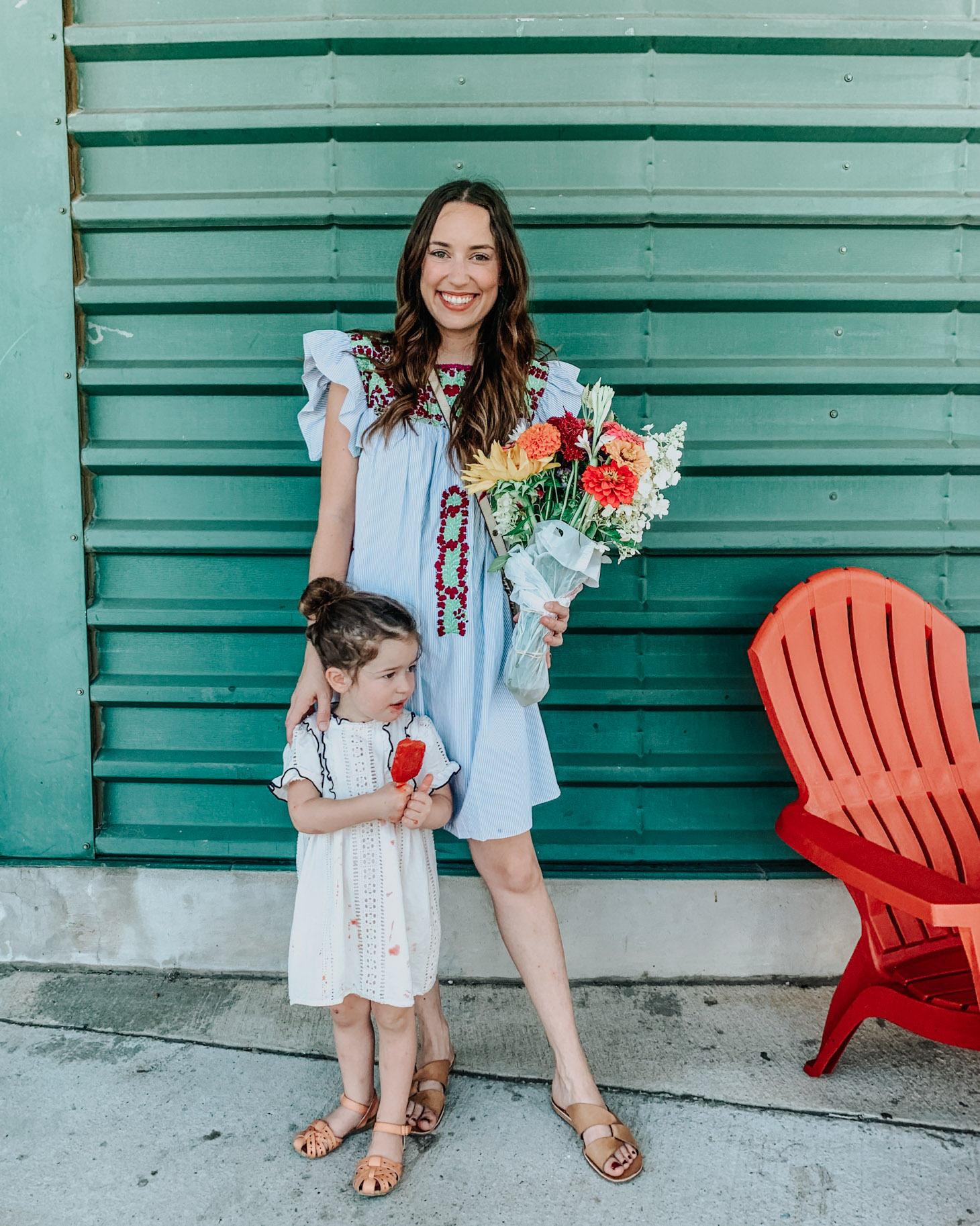 5 Dress Designers to Take Note Of this Summer by popular Memphis fashion blog, Lone Star Looking Glass: image of a woman wearing a Mi Golondrina floral embroidered dress and standing in front of a green industrial wall with her daughter who is wearing a white eyelet dress and eating a red popsicle. 