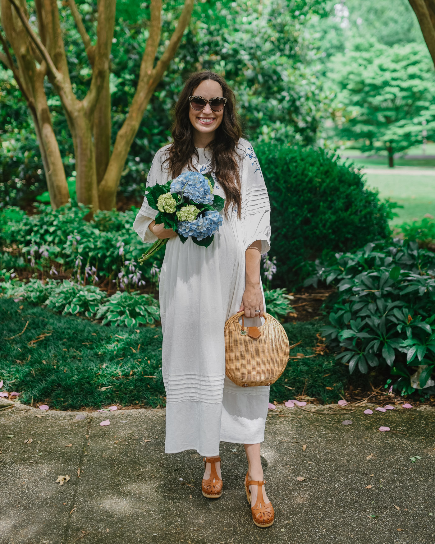 Meadows Dresses by popular Memphis fashion blog, Lone Star Looking Glass: image of a woman standing outside while holding a bouquet of hydrangeas and wearing a Meadows Azelea dress, brown clog sandals, and carrying a wicker handbag. 