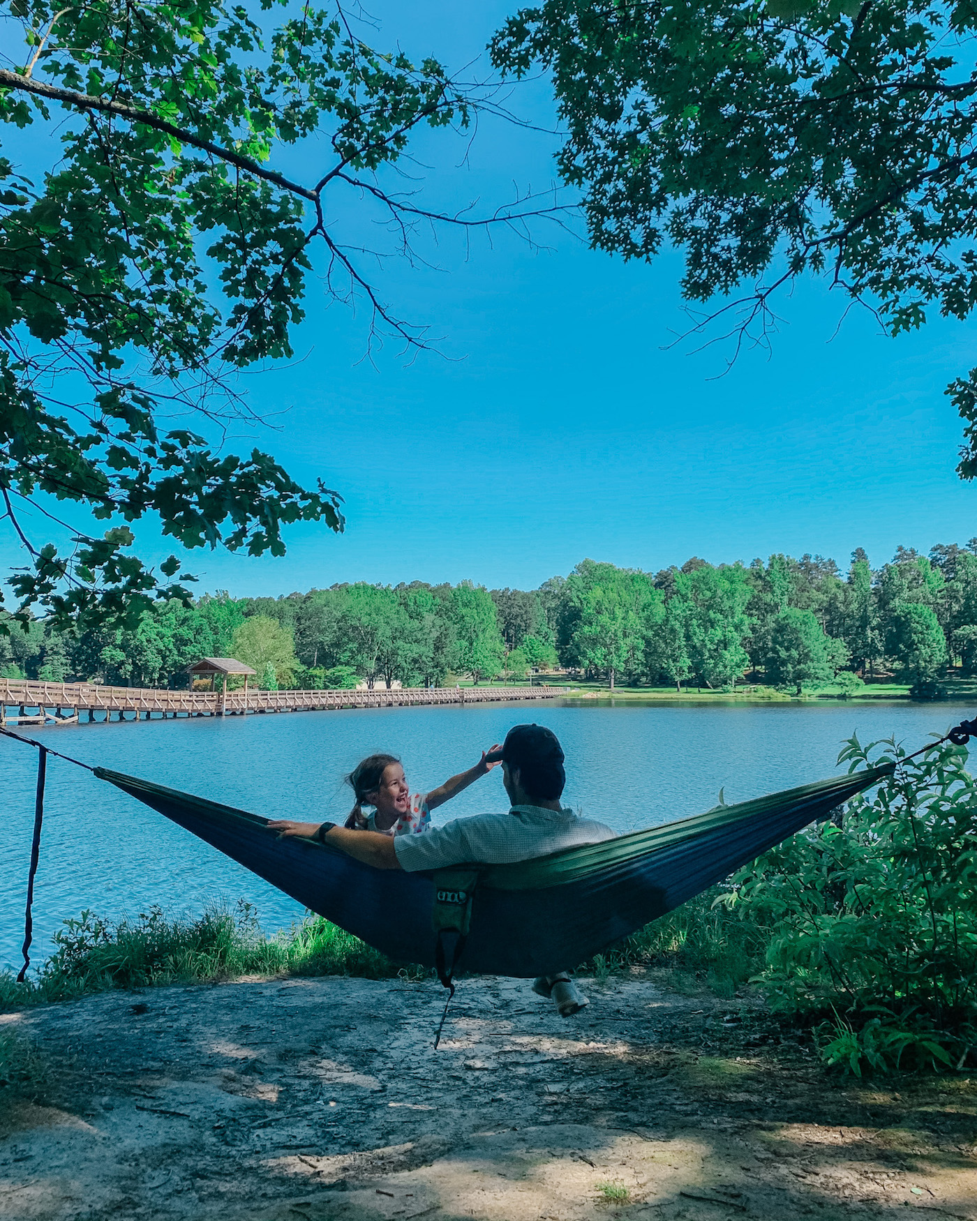 Father's Day Gift Ideas by popular Memphis lifestyle blog, Lone Star Looking Glass: image of a dad and his young daughter sitting in a hammock in front of a lake. 