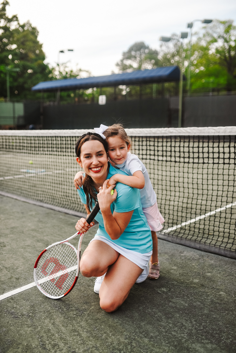 Tennis Outfits by popular Memphis fashion blog, Lone Star Looking Glass: image of a mom kneeling on a tennis court and holding a tennis racket and tennis ball while her young daughter hugs her from behind and they're both wearing tennis outfits. 