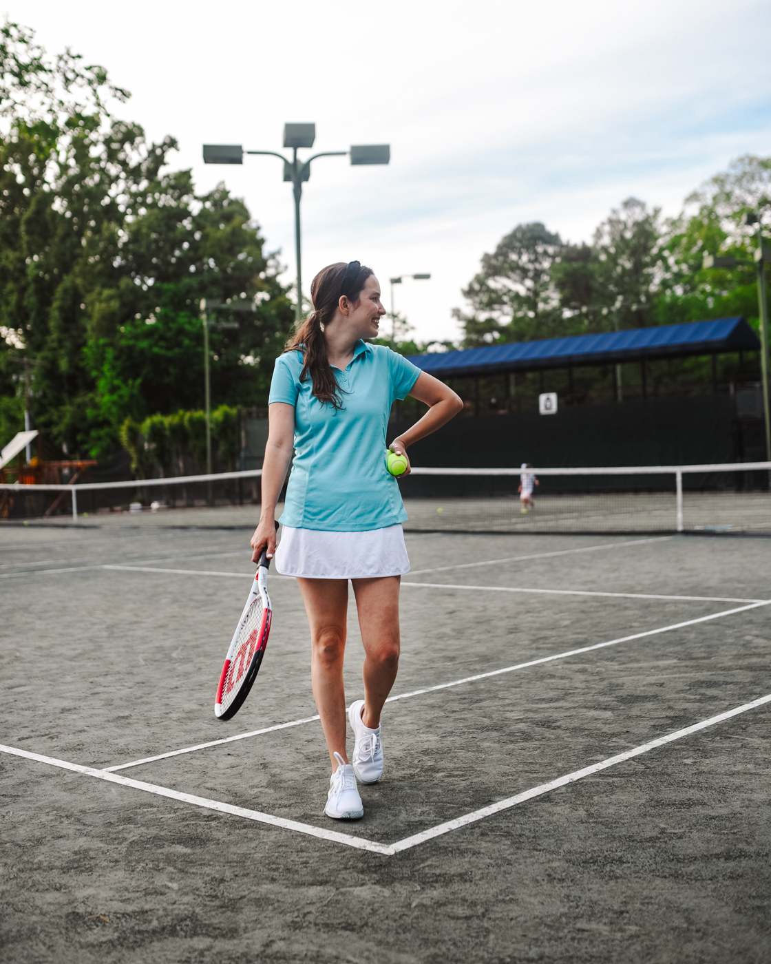 Tennis Outfits by popular Memphis fashion blog, Lone Star Looking Glass: image of a woman standing on a tennis court and holding a tennis racket and tennis ball and wearing a white tennis skirt, blue tennis polo and white sneakers. 