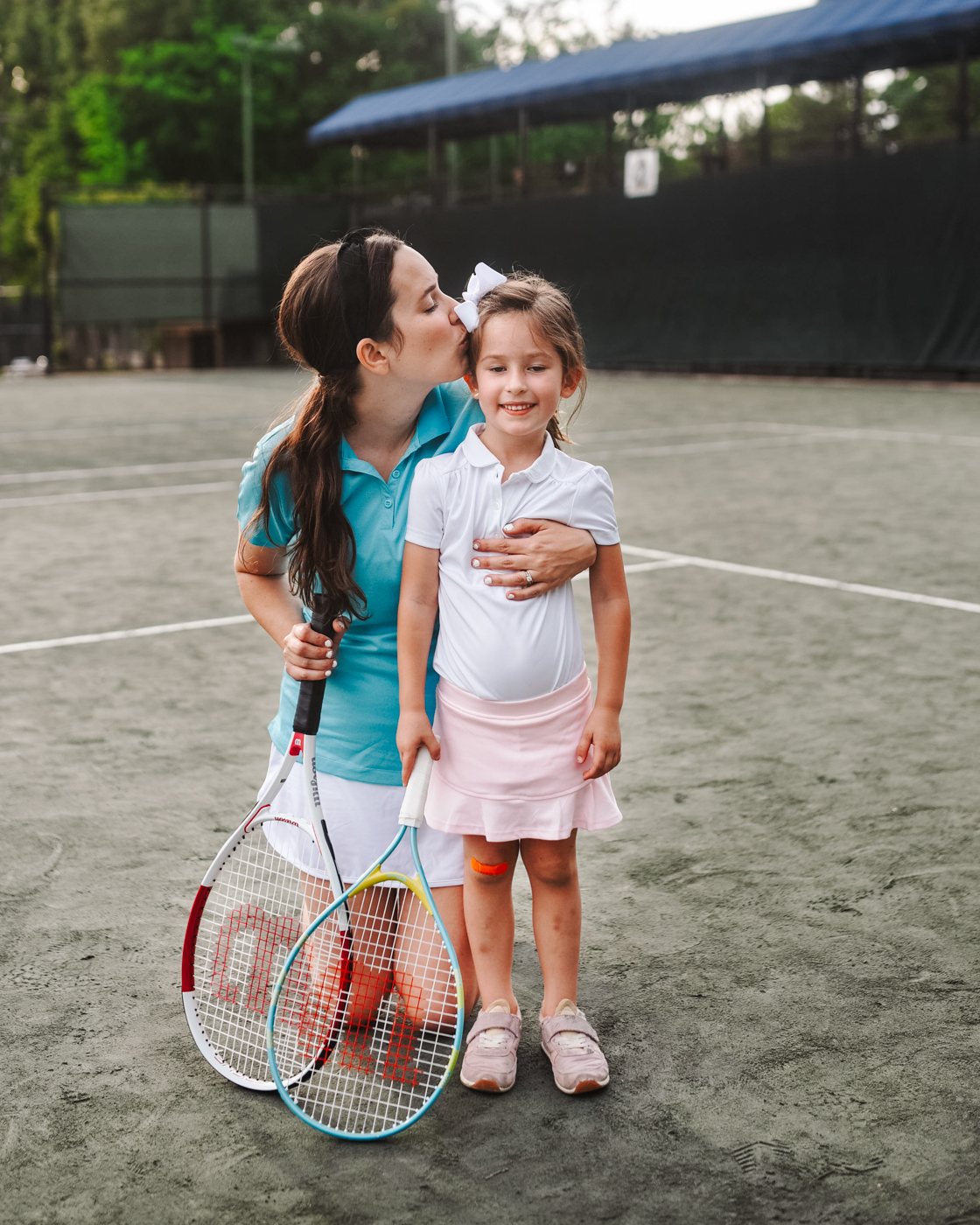 Tennis Outfits by popular Memphis fashion blog, Lone Star Looking Glass: image of a mom kissing her daughter on the cheek as they stand on a tennis court and hold tennis rackets and wearing tennis skirts, sneakers, and tennis polos. 