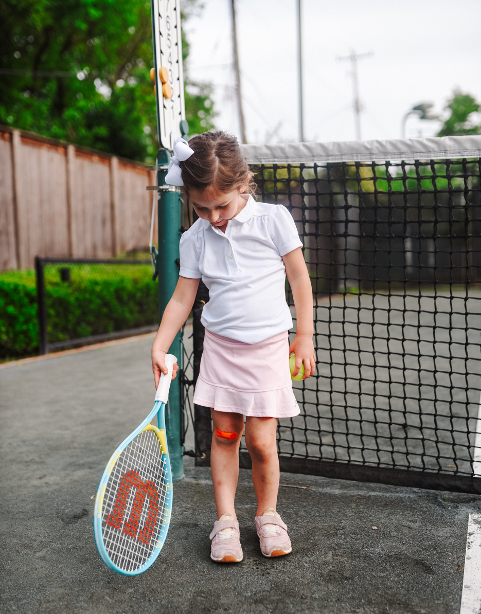 Tennis Outfits by popular Memphis fashion blog, Lone Star Looking Glass: image of a young girl standing on a tennis court and holding a tennis racket while wearing a pink tennis skirt, white polo tennis shirt and white bow in her hair. 