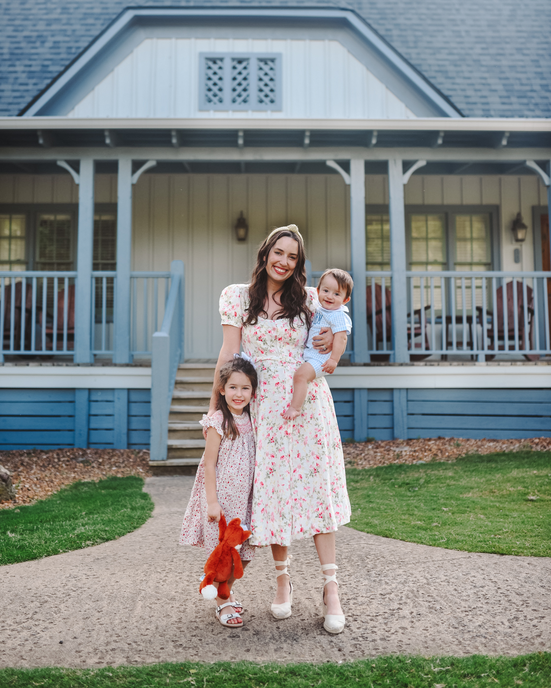Barnsley Resort by popular Memphis travel blog, Lone Star Looking Glass: image of a mom and standing next to her daughter and holding her young son and standing in front of a White House with a blue porch and wearing a floral print midi dress, lace up espadrilles, knot headband, smock floral print dress, white sandals, and blue smock romper. 