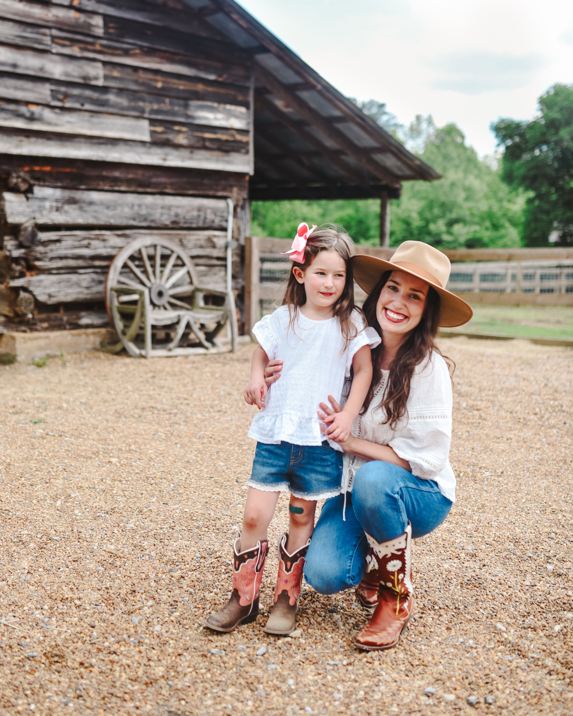 Barnsley Resort by popular Memphis travel blog, Lone Star Looking Glass: image of a mom and her young daughter wearing white shirts, lace trim jean shorts, jeans, cowboy boots, strap rancher hat and pink bow and standing next to a old wood barn. 