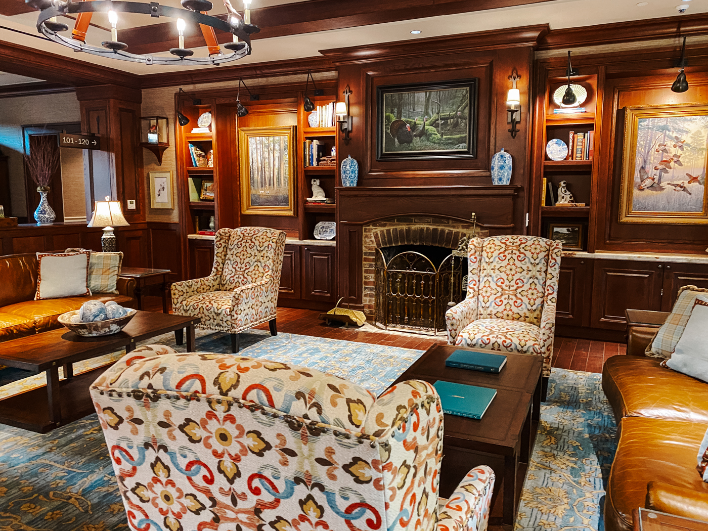 Barnsley Resort by popular Memphis travel blog, Lone Star Looking Glass: image of a sitting room with a red brick fireplace, dark wood trim, dark wood built in shelves, leather couches, blue and cream area rug, oil paintings of ducks in gold frames, and floral print arm chairs. 