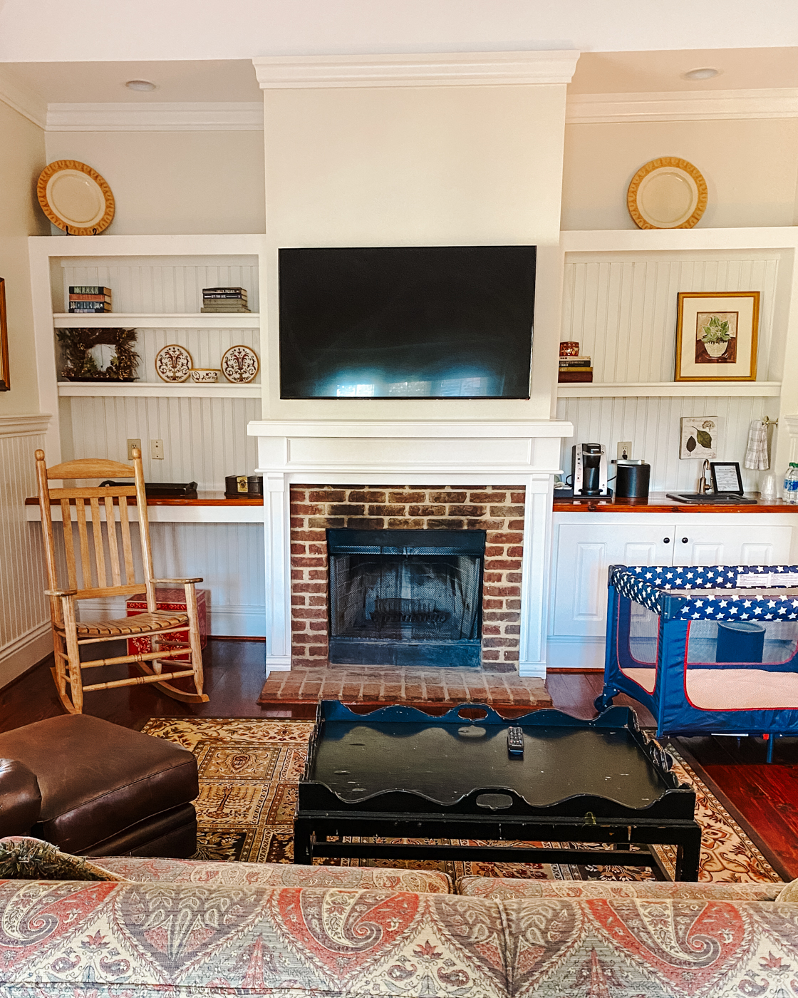 Barnsley Resort by popular Memphis travel blog, Lone Star Looking Glass: image of a room with a red brick fireplace with a white mantle and a flat screen t.v. hanging above, black coffee table, built in white bookshelves, portable crib, rocking chair, cherry wood floors, and couch with red, brown and cream paisley fabric. 