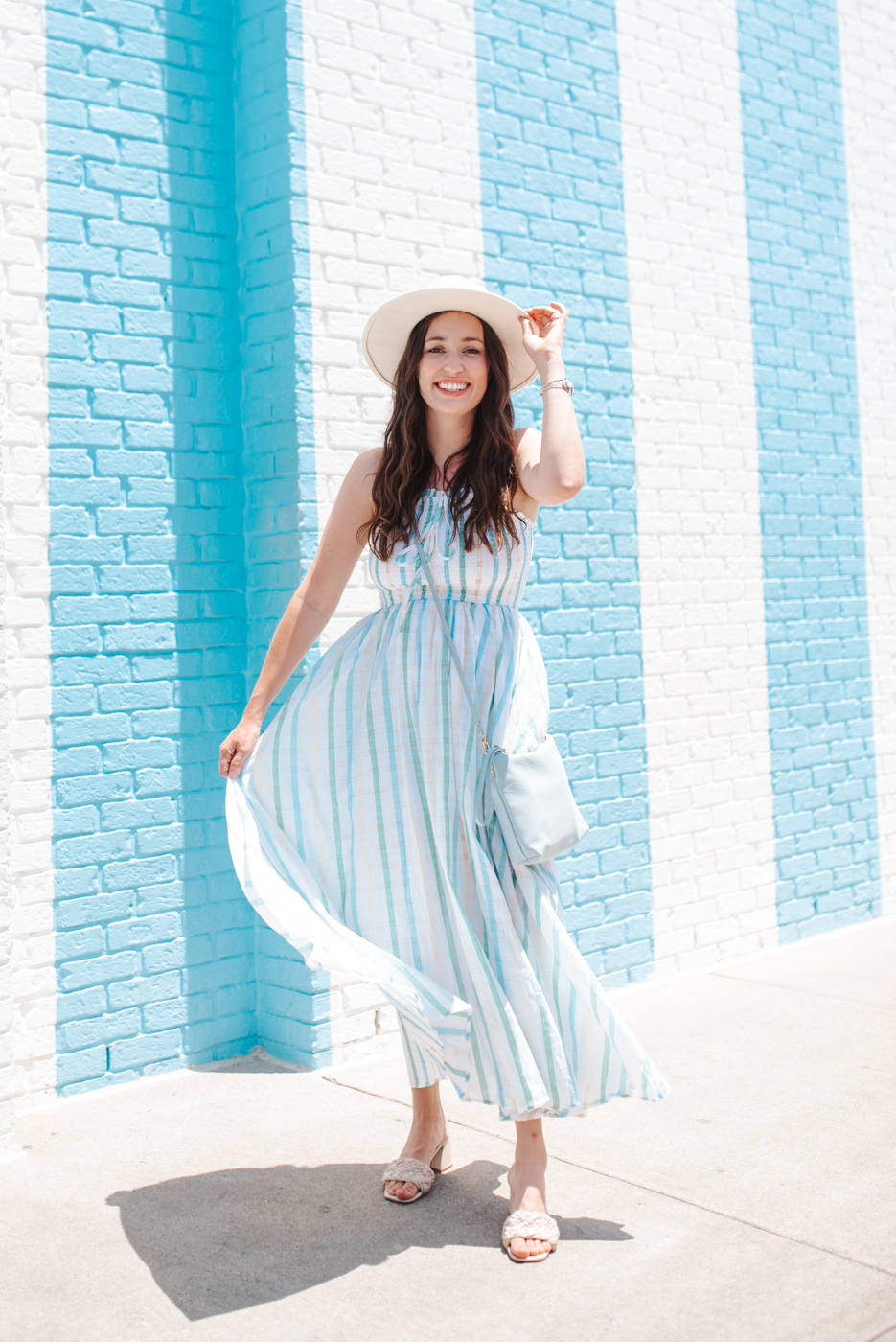 Gingham Dress by popular Memphis fashion blog, Lone Star Looking Glass: image of a woman standing next to a blue and white stripe brick wall and wearing a white straw hat, white slide sandals, and a blue and green gingham dress. 