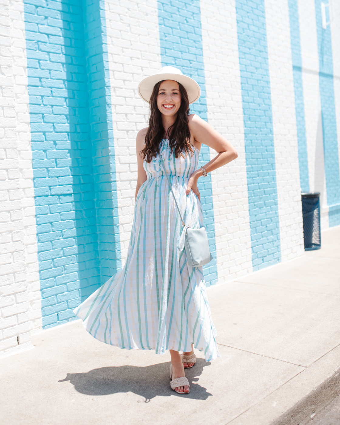 Gingham Dress by popular Memphis fashion blog, Lone Star Looking Glass: image of a woman standing next to a blue and white stripe brick wall and wearing a white straw hat, white slide sandals, and a blue and green gingham dress. 