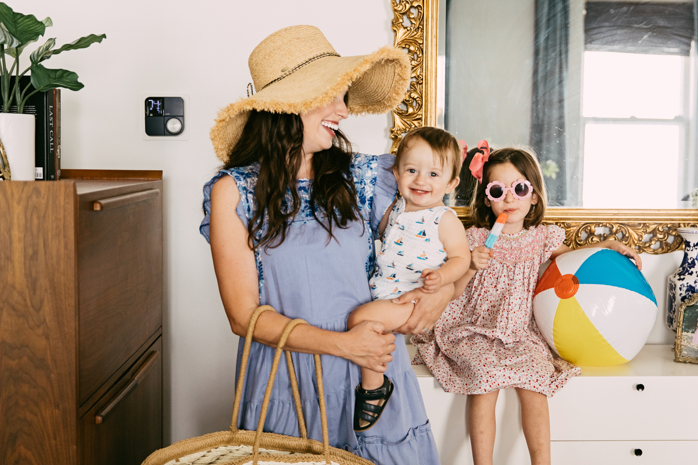 Lux Kono Smart Thermostat by popular Memphis lifestyle blog: image of a mom wearing a straw floppy brim sunhat and blue chambray dress and standing next to her two kids as they sit on a modern white dresser in front of a gilded gold frame mirror. 