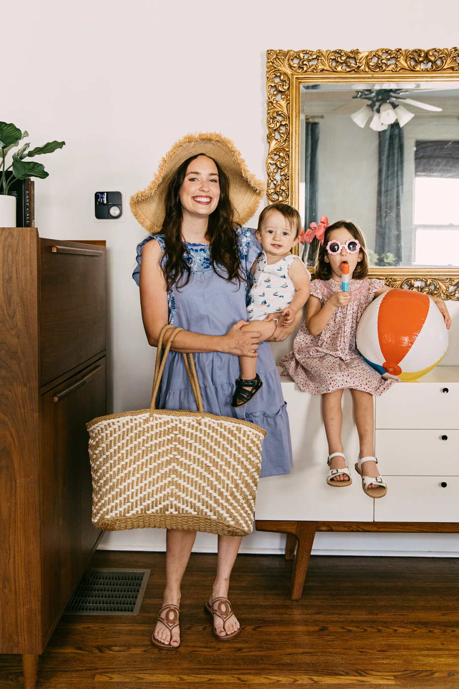 Lux Kono Smart Thermostat by popular Memphis lifestyle blog: image of a mom wearing a straw floppy brim sunhat and blue chambray dress and standing next to her two kids as they sit on a modern white dresser in front of a gilded gold frame mirror. 